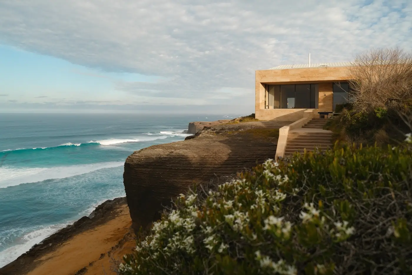 A sandstone and glass house stepping down a limestone cliff above a turquoise bay