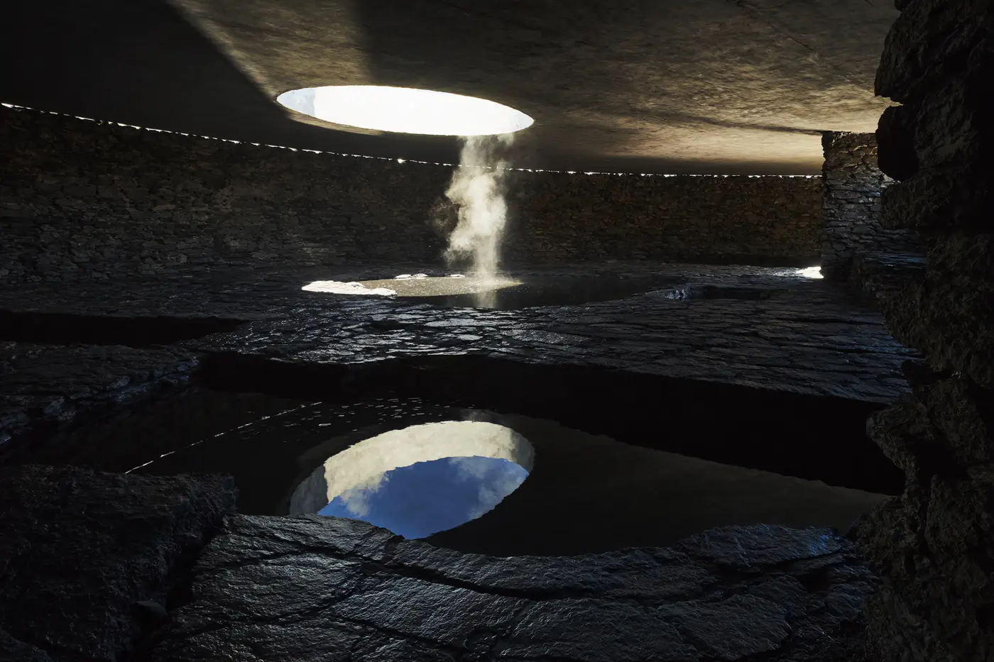 Dark basalt thermal pools under a concrete oculus with a shaft of sunlight through steam